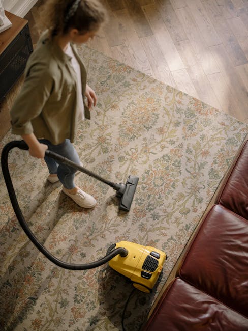 A young boy using a vacuum cleaner to perform deep surface cleaning on a beige sofa in a well-lit living room. The room features a hardwood floor partially covered by an orange and gray rug, with a co
